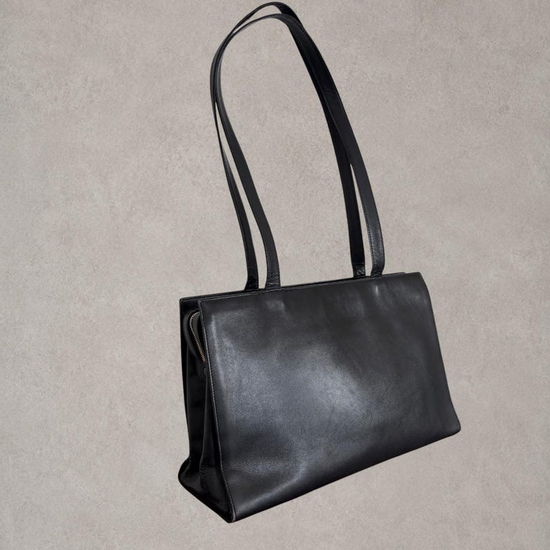 Puede incluir: Un bolso de hombro de cuero negro con silueta rectangular, que cuenta con dos correas largas y finas y un cierre de cremallera lateral. El bolso tiene un acabado liso y mate y una base estructurada, ofreciendo un dise&ntilde;o limpio y funcional.