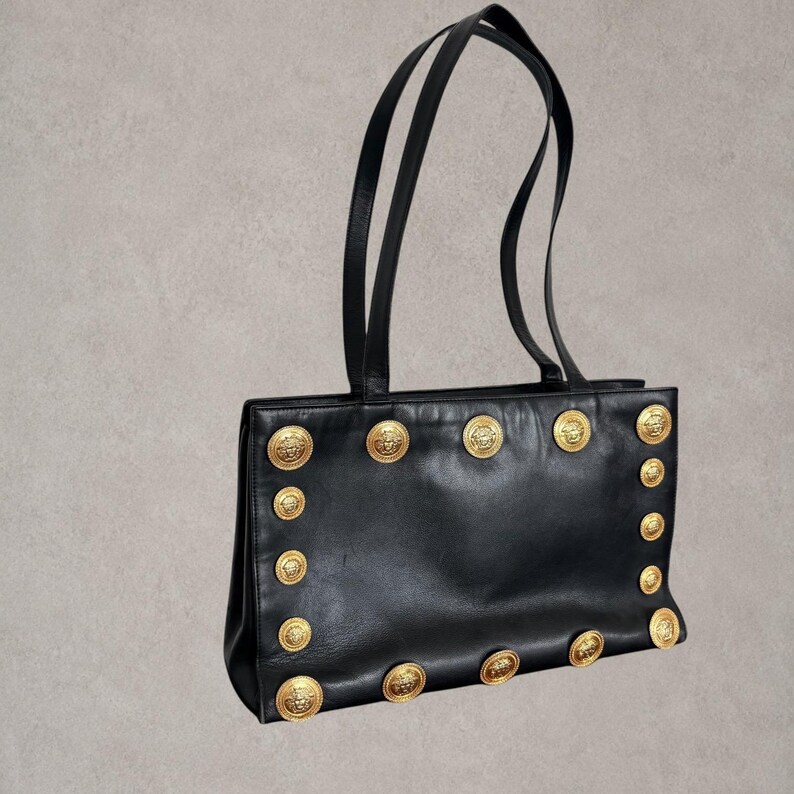 Puede incluir: Un bolso tote de cuero negro con dos asas largas para el hombro y una silueta rectangular. La parte frontal del bolso est&aacute; decorada con una serie de botones met&aacute;licos circulares en tono dorado, cada uno con un relieve de cabeza de Medusa.