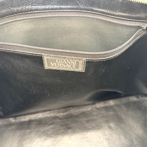 Puede incluir: El interior de un bolso de cuero negro con un bolsillo con cremallera en el borde superior. Una etiqueta de tela rectangular est&aacute; cosida debajo de la cremallera, mostrando el texto GIANNI VERSACE en letras may&uacute;sculas blancas.