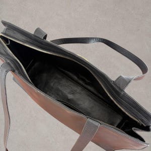 Puede incluir: Un bolso tote con dise&ntilde;o bicolor que combina un exterior marr&oacute;n oscuro con ribetes, asas y forro interior de cuero negro. El bolso cuenta con un cierre de cremallera superior con riel met&aacute;lico y un amplio compartimento principal abierto.