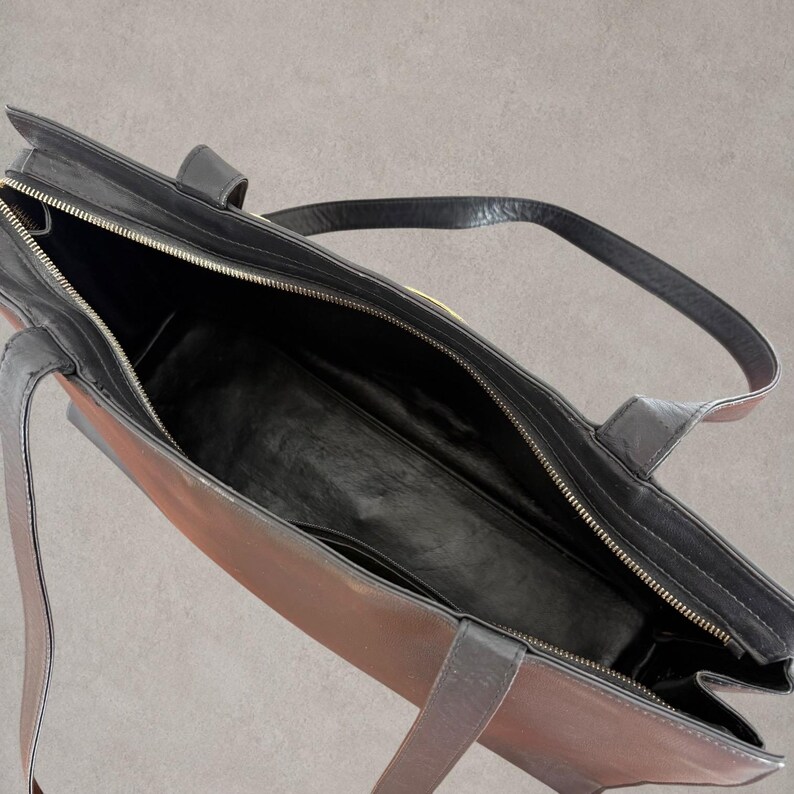Puede incluir: Un bolso tote con dise&ntilde;o bicolor que combina un exterior marr&oacute;n oscuro con ribetes, asas y forro interior de cuero negro. El bolso cuenta con un cierre de cremallera superior con riel met&aacute;lico y un amplio compartimento principal abierto.