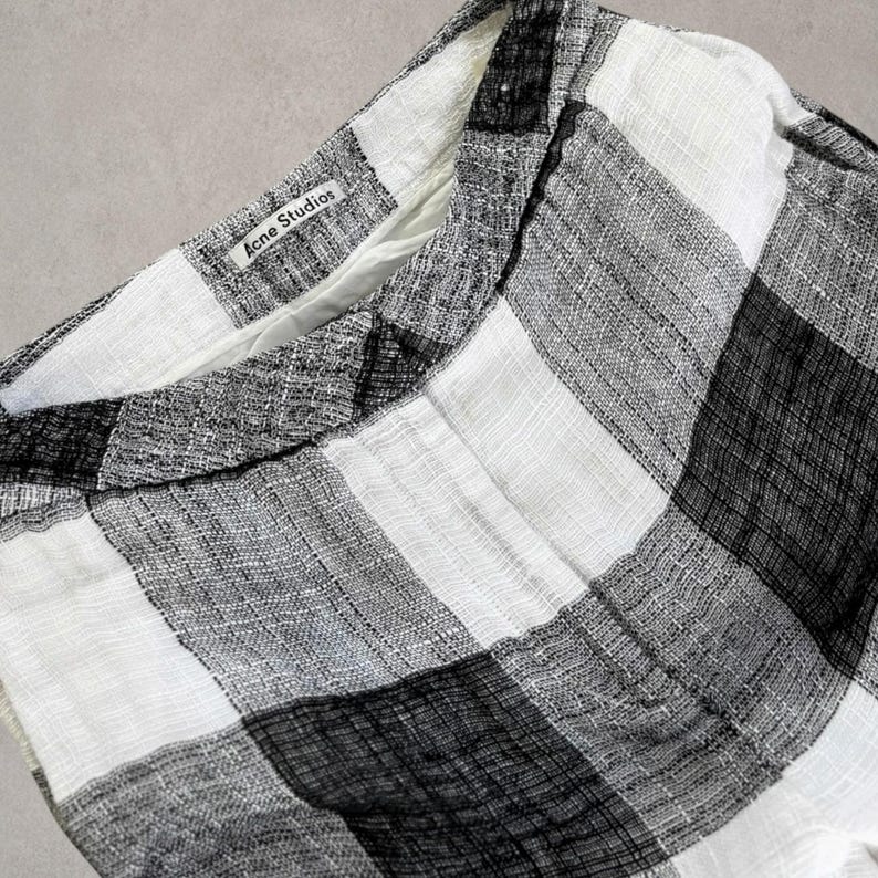 Puede incluir: Un pantal&oacute;n con un patr&oacute;n de cuadros en blanco y negro con un acabado de tela tejida texturizada. La cintura incluye una etiqueta interior blanca con el texto negro Acne Studios impreso, visible en el borde superior de la prenda.