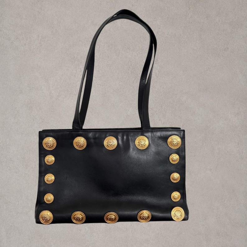 Puede incluir: Un bolso tote de cuero negro con dos correas para el hombro y un cuerpo rectangular. La parte frontal est&aacute; decorada con un borde de medallones met&aacute;licos circulares en tono dorado, cada uno grabado con un motivo detallado de Medusa.