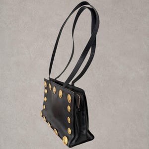Puede incluir: Un bolso de hombro negro con dos correas largas y finas y un cuerpo rectangular. El panel frontal est&aacute; decorado con una serie de medallones met&aacute;licos dorados dispuestos a lo largo de los bordes. Se observa un cierre de cremallera met&aacute;lico en el lateral.
