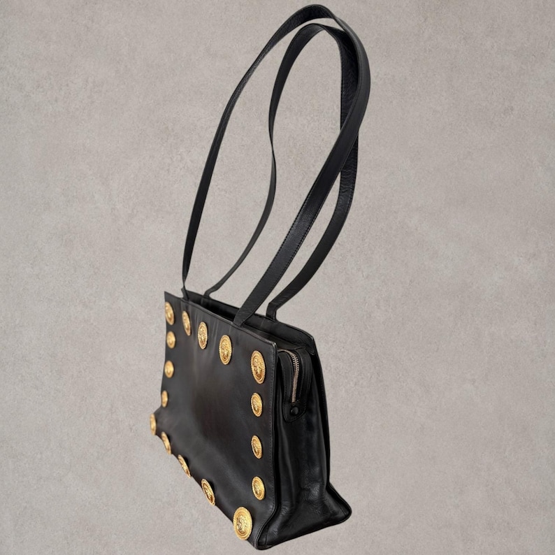 Puede incluir: Un bolso de hombro negro con dos correas largas y finas y un cuerpo rectangular. El panel frontal est&aacute; decorado con una serie de medallones met&aacute;licos dorados dispuestos a lo largo de los bordes. Se observa un cierre de cremallera met&aacute;lico en el lateral.