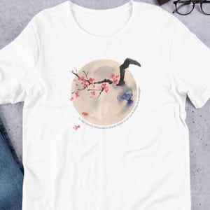 May include: White t-shirt featuring a watercolor design of a full moon with cherry blossom branches and the text "In the cherry blossom's shade there is no such thing as a stranger". The design includes red, black, and blue accents.