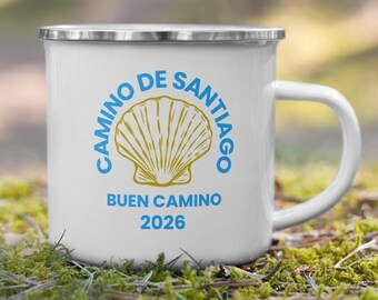 May include: White enamel mug with a silver rim and handle. The mug features a blue and gold design with the words "Camino de Santiago" and "Buen Camino 2026" surrounding a scallop shell graphic. The mug is sitting on a bed of green moss.