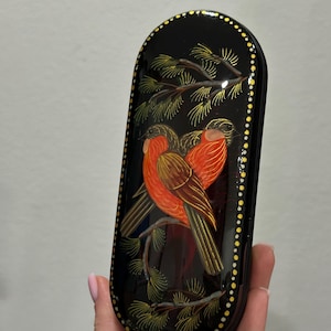 May include: Black, oval-shaped decorative box with a painted design of two red birds perched on a pine branch. The box has a glossy finish and a border of gold dots. The birds have brown and red feathers.
