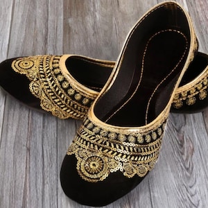 Puede incluir: Un par de bailarinas negras con intrincados bordados dorados y detalles de lentejuelas. Los zapatos tienen una punta redondeada y un ribete dorado. El diseño presenta patrones florales y geométricos, creando un aspecto elegante y ornamentado.