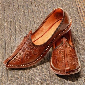 Puede incluir: Un par de zapatos sin cordones de cuero marrón con intrincados patrones florales en relieve. Los zapatos tienen una punta puntiaguda y una suela cosida con hilo blanco. El interior es de un tono marrón más claro.