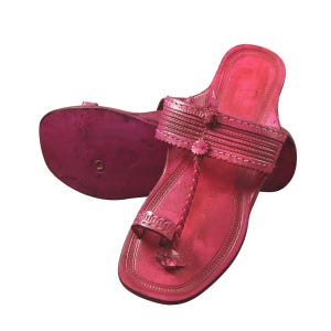 Könnte beinhalten: Ein Paar leuchtend pinkfarbene Ledersandalen. Die Sandalen haben eine Zehenschlaufe, geflochtene Riemen und dekorative Nähte. Die Sohlen sind in einem dunkleren Rosaton gehalten. Die Sandalen sind auf Komfort und Stil ausgelegt.