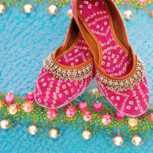 Puede incluir: Un par de zapatos rosas bordados sin cordones con un patrón bandhani. Los zapatos tienen un ribete de cuentas decorativo y una suela de cuero marrón. El fondo es una superficie azul claro con detalles florales y perlas.