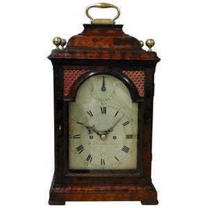 May include: An antique wooden clock with a handle and brass details. The clock face features Roman numerals and the words "STRIKE" and "SILENT". The clock is brown with red patterned detailing, and is approximately 30 cm tall.
