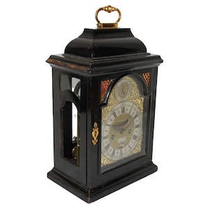 Puede incluir: Reloj de madera negro con asa dorada y detalles dorados decorativos. La esfera del reloj es dorada con números romanos negros. El reloj tiene una parte delantera de cristal y un péndulo visible en el interior. El reloj es rectangular.