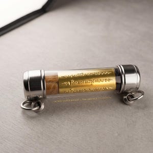 以下が含まれることがあります： 透明なチューブの中に金色の刻印が入った円筒形のお守り。端は銀色の金属で覆われ、小さなリングが付いています。「Sacred Takrut Amulet | Thai Heritage Collection」の文字が見えます。