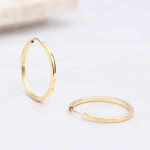 May include: Two gold hoop earrings on a white surface. The earrings are circular and have a smooth, polished finish. In the background, a decorative arrangement of flowers and a rectangular stone.