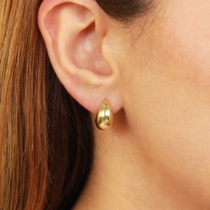 May include: A close-up of a gold teardrop earring on an ear. The earring is smooth and shiny, reflecting light. The ear is set against a background of reddish-brown hair and skin.