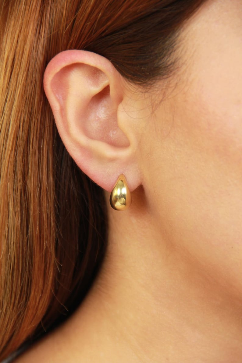 May include: A close-up of a gold teardrop earring on an ear. The earring is smooth and shiny, reflecting light. The ear is set against a background of reddish-brown hair and skin.