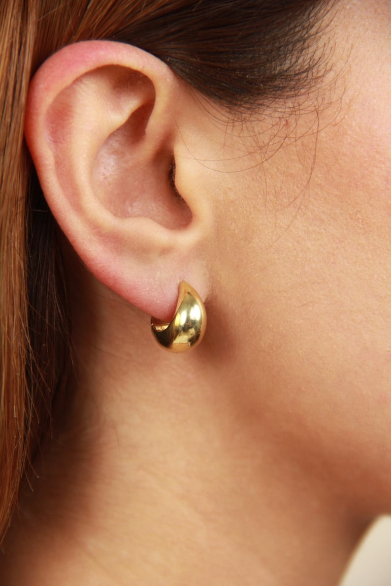 May include: A close-up of a gold-colored, teardrop-shaped earring. The earring is a small, polished hoop design, reflecting light. The earring is worn on a person's ear.