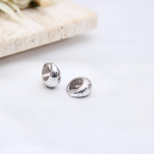 May include: Two silver-colored, teardrop-shaped hoop earrings. The earrings are smooth and shiny, reflecting light. They are displayed on a white surface, with a light-colored rectangular block and floral accents in the background. Jewelry.