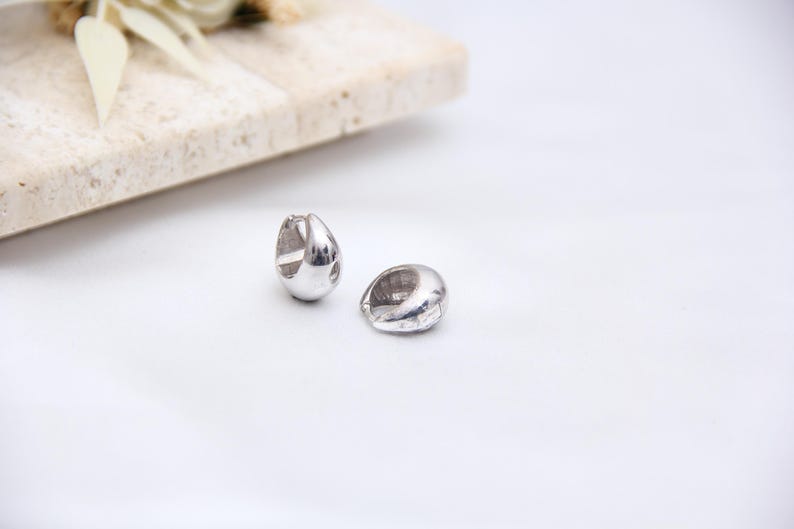 May include: Two silver-colored, teardrop-shaped hoop earrings. The earrings are smooth and shiny, reflecting light. They are displayed on a white surface, with a light-colored rectangular block and floral accents in the background. Jewelry.