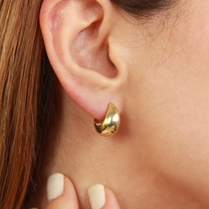 May include: A close-up of a gold-colored, teardrop-shaped hoop earring. The earring is worn on an ear, with the skin tone appearing fair. The earring has a smooth, polished surface and a rounded shape.