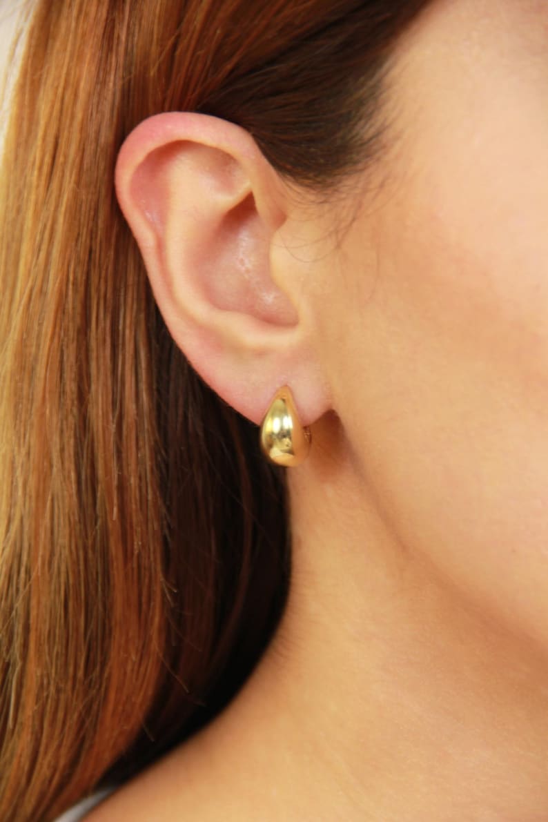 May include: A close-up of a gold teardrop earring. The earring is a simple, polished design with a smooth, reflective surface. The earring is worn on an ear, with the person's hair and skin visible in the background.