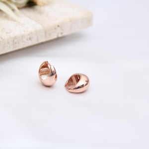May include: Two rose gold teardrop-shaped earrings on a white surface. The earrings have a smooth, polished finish and a small opening on one side. A light-colored stone slab and dried flowers are in the background.