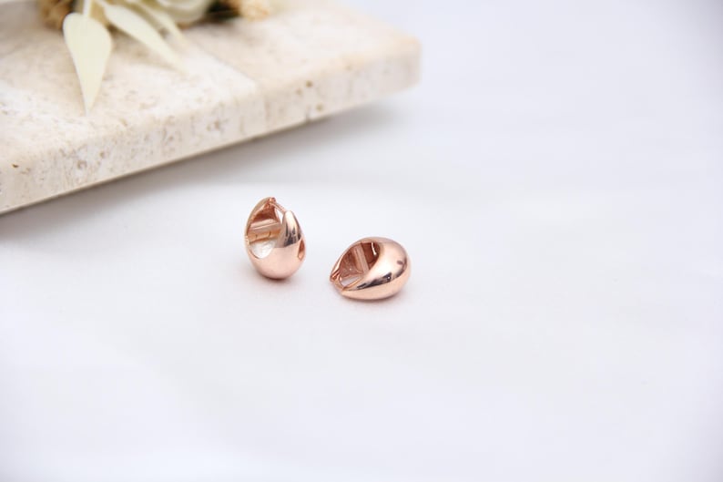May include: Two rose gold teardrop-shaped earrings on a white surface. The earrings have a smooth, polished finish and a small opening on one side. A light-colored stone slab and dried flowers are in the background.