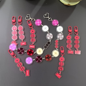 Könnte beinhalten: Eine Sammlung von Acryl-Schlüsselanhängern mit roter Hardware. Die Schlüsselanhänger weisen eine Vielzahl von Designs auf, darunter kreisförmige Scheiben in Rosa-, Lila- und Rottönen sowie rechteckige Namensschilder. Einige Schlüsselanhänger haben herzförmige Verschlüsse.