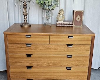 May include: A light brown wooden dresser with six drawers and black handles. The top of the dresser displays a decorative candelabra, a vase of greenery, a small statue, and books. The dresser is set against a white curtain.