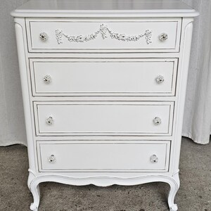May include: A white painted chest of drawers with four drawers and crystal knobs. The top drawer features a decorative floral design. The chest has curved legs and a distressed finish, giving it a vintage look.