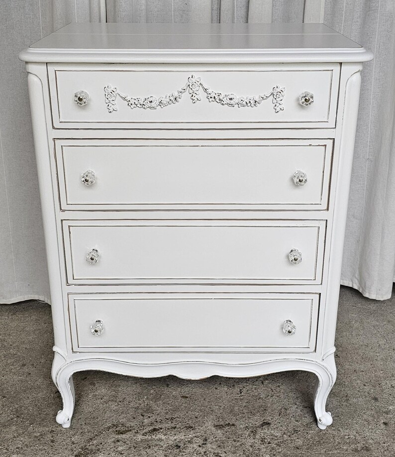 May include: A white painted chest of drawers with four drawers and crystal knobs. The top drawer features a decorative floral design. The chest has curved legs and a distressed finish, giving it a vintage look.