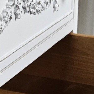 May include: White dresser with an open drawer. The dresser features ornate silver-colored floral detailing and a crystal-like drawer pull. The inside of the drawer is made of wood. The dresser is a classic piece of furniture.