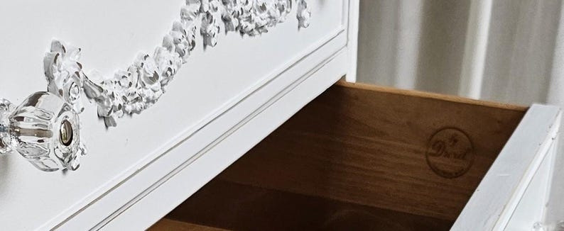 May include: White dresser with an open drawer. The dresser features ornate silver-colored floral detailing and a crystal-like drawer pull. The inside of the drawer is made of wood. The dresser is a classic piece of furniture.
