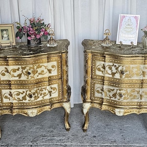 May include: Two antique bedside tables with a gold and cream finish. Each table has two drawers and ornate gold detailing. The tops are decorated with small framed pictures, flowers, and decorative objects. The tables have curved legs and a distressed look.