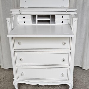May include: A white antique desk with a drop-down writing surface and three drawers. The desk features crystal knobs and small drawers and compartments. The desk has a curved design and is painted white.