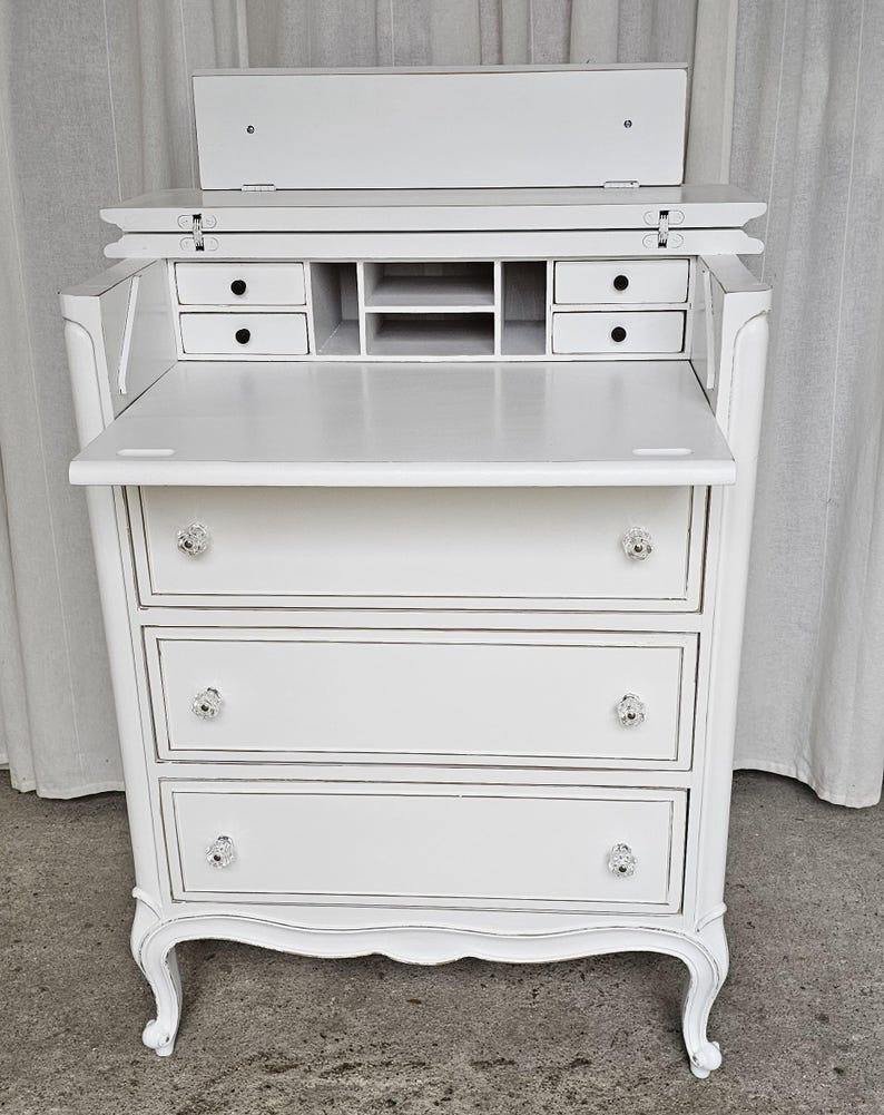 May include: A white antique desk with a drop-down writing surface and three drawers. The desk features crystal knobs and small drawers and compartments. The desk has a curved design and is painted white.