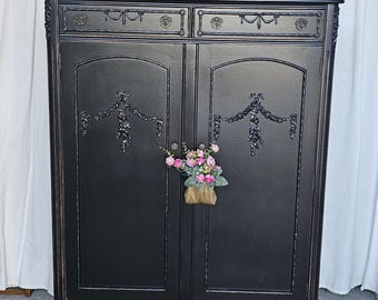 May include: A black antique cabinet with ornate detailing, featuring two doors and two drawers. The cabinet is adorned with floral accents and decorative carvings. A small bouquet of pink flowers hangs from the doors. A framed picture and a candle holder sit on top.