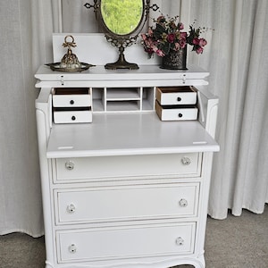 May include: A white antique desk with a fold-down writing surface, three drawers, and crystal knobs. It features a decorative mirror, a small tray, and a vase of flowers. The desk has a vintage, elegant aesthetic.