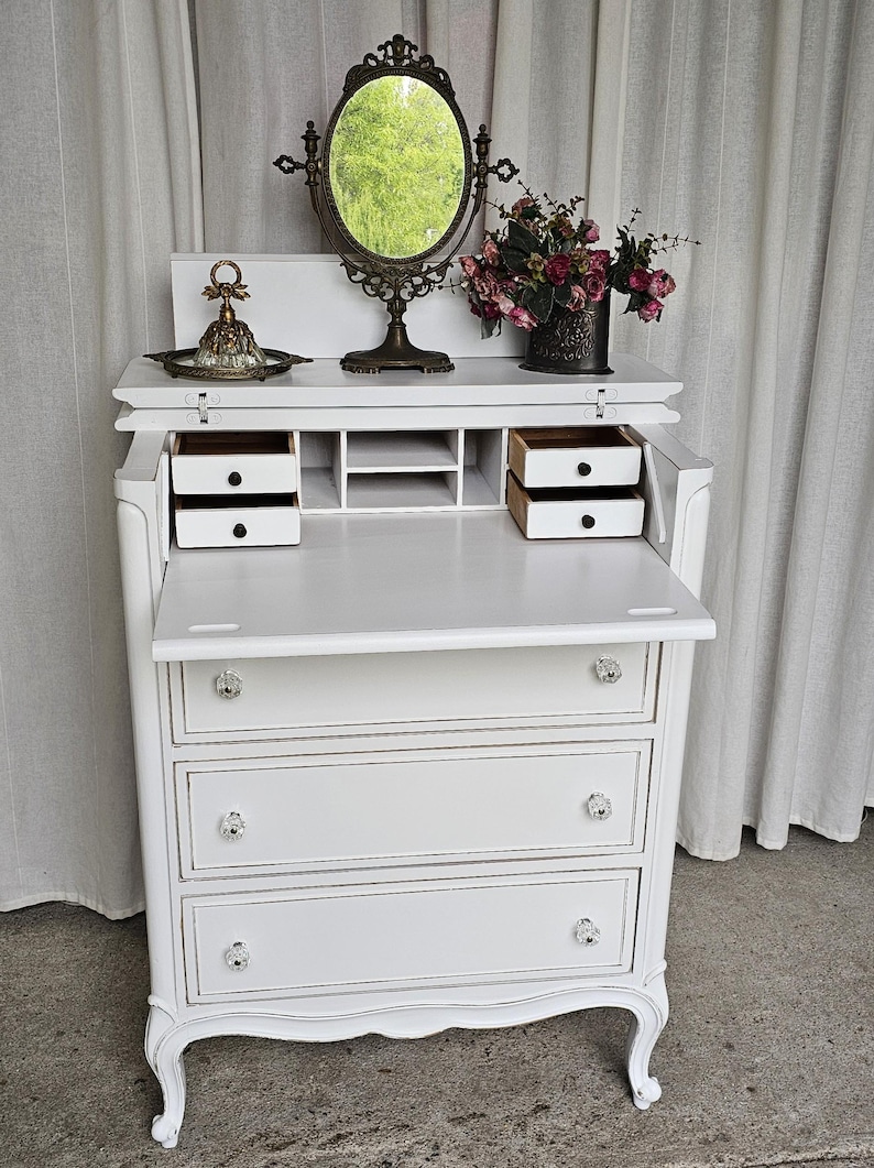 May include: A white antique desk with a fold-down writing surface, three drawers, and crystal knobs. It features a decorative mirror, a small tray, and a vase of flowers. The desk has a vintage, elegant aesthetic.
