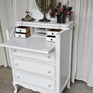 May include: White antique secretary desk with three drawers, crystal knobs, and a drop-down writing surface. The desk features a decorative mirror, a small vase of flowers, and two small drawers. The desk is a classic piece of furniture.