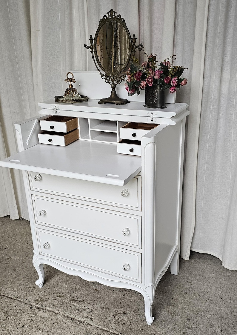 May include: White antique secretary desk with three drawers, crystal knobs, and a drop-down writing surface. The desk features a decorative mirror, a small vase of flowers, and two small drawers. The desk is a classic piece of furniture.