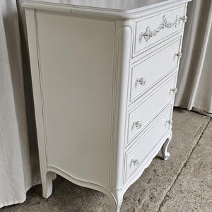 May include: A white painted chest of drawers with five drawers and crystal knobs. The chest has a decorative floral design on the top drawer and curved legs. The drawers are outlined with a gray trim.