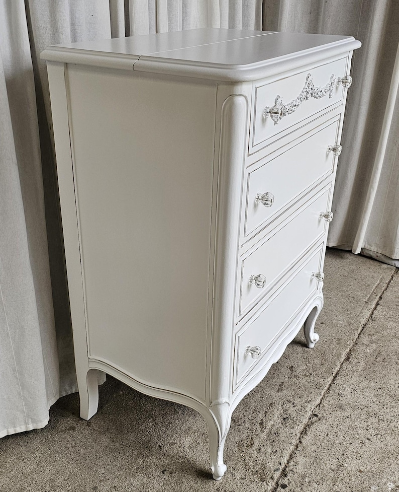 May include: A white painted chest of drawers with five drawers and crystal knobs. The chest has a decorative floral design on the top drawer and curved legs. The drawers are outlined with a gray trim.