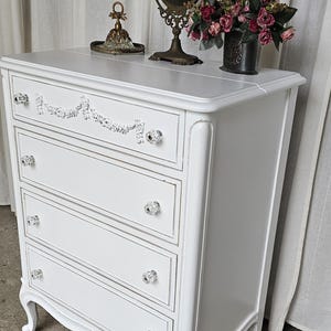 May include: A white painted dresser with four drawers, crystal knobs, and decorative trim. The dresser has a curved front and legs. A vintage mirror, flowers, and decorative items sit on top.