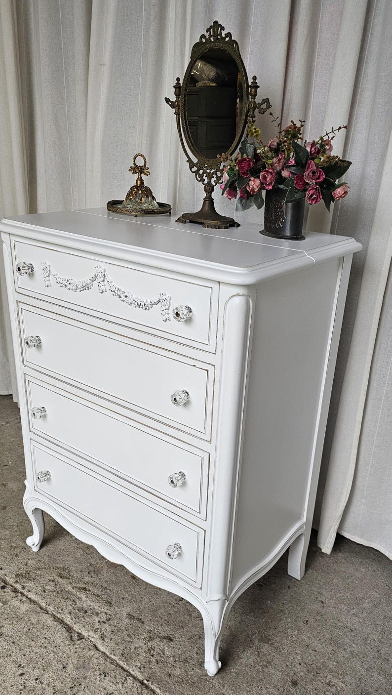 May include: A white painted dresser with four drawers, crystal knobs, and decorative trim. The dresser has a curved front and legs. A vintage mirror, flowers, and decorative items sit on top.