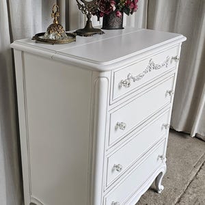May include: White painted dresser with four drawers, crystal knobs, and ornate detailing. A decorative mirror, flowers, and a perfume bottle sit on top. The dresser has curved legs and a classic design.
