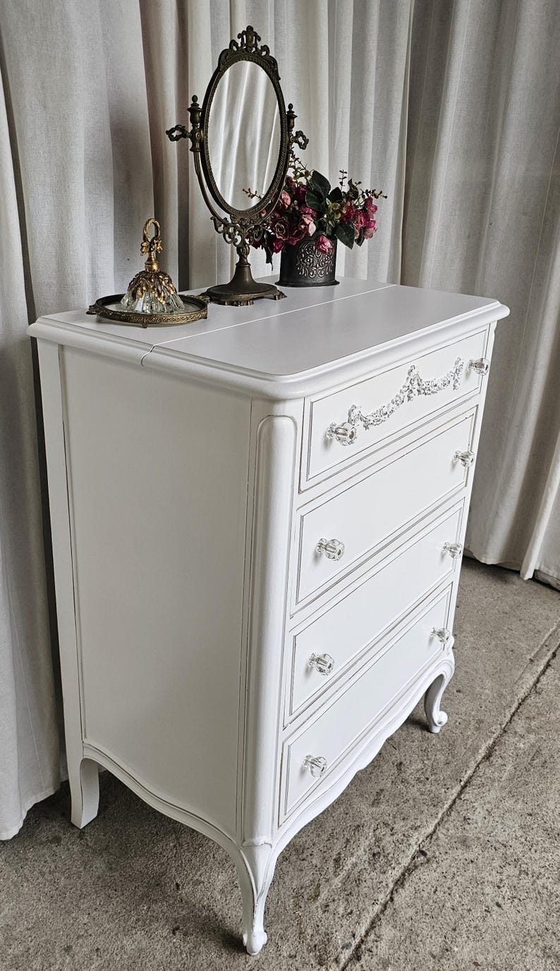 May include: White painted dresser with four drawers, crystal knobs, and ornate detailing. A decorative mirror, flowers, and a perfume bottle sit on top. The dresser has curved legs and a classic design.