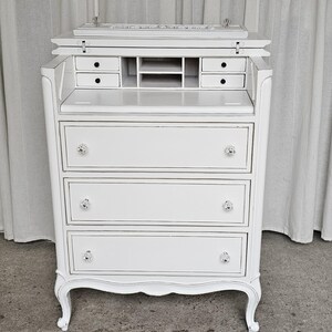 May include: A white, vintage-style secretary desk with a drop-down writing surface. The desk features three drawers with crystal knobs, small interior drawers, and a storage compartment. The desk has a curved base and ornate detailing, and is painted white with silver trim.